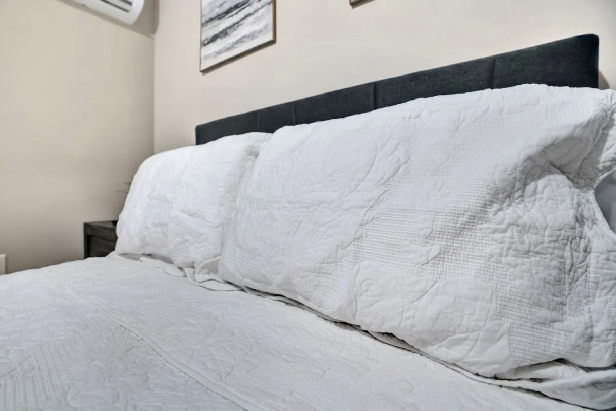 A bedroom centered on a plush bed with white quilted bedding, a dark headboard, and tasteful wall art. The overall feel is one of serenity and coziness, inviting relaxation and restful sleep.