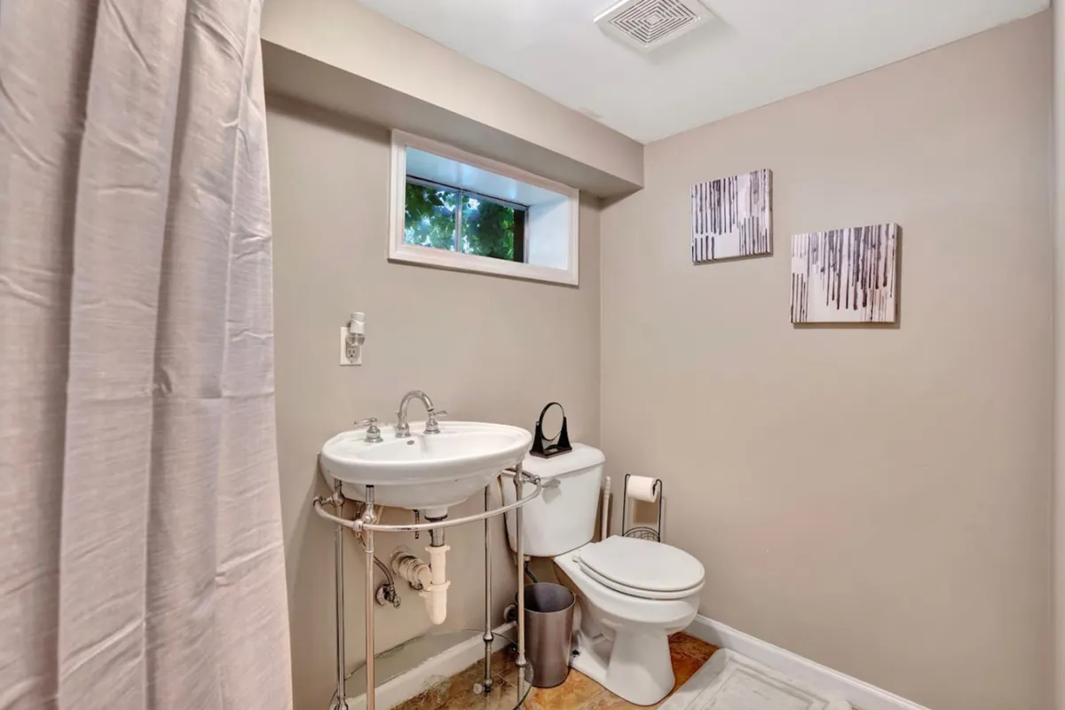 A serene bathroom scene with a stylish sink, modern toilet, framed artwork, and a window allowing natural light to brighten the space. The setup feels clean, fresh, and thoughtfully designed for comfort.