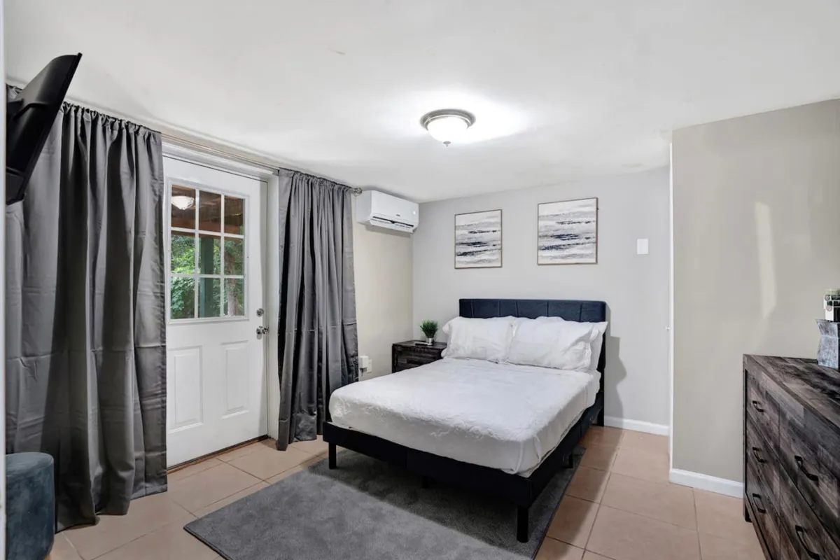 An inviting bedroom featuring a neatly made bed, soft curtains framing natural light, a nightstand with a lamp, and calming artwork on the wall. The ambiance exudes serenity and peace.