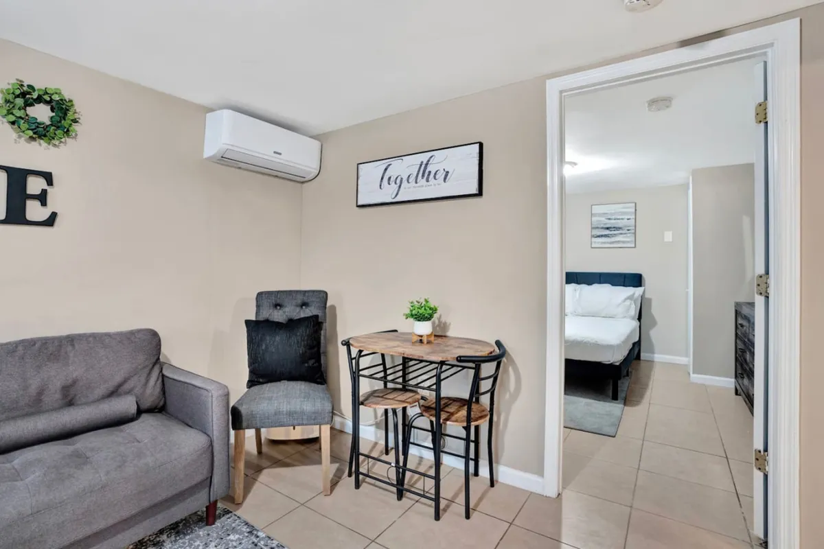 This image features a cozy living area with a comfortable sofa, a small dining table perfect for intimate meals, decorative wall art that adds a personal touch, and an air conditioning unit to ensure comfort. The soft lighting and inviting arrangement suggest a welcoming space to unwind and connect.