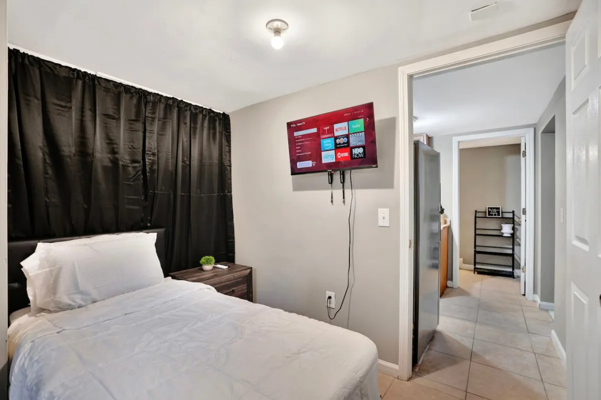 A cozy bedroom with a bed dressed in crisp white bedding, dark curtains for privacy, and a TV mounted on the wall. The room feels peaceful and comfortable, ideal for relaxation or unwinding with a favorite show.
