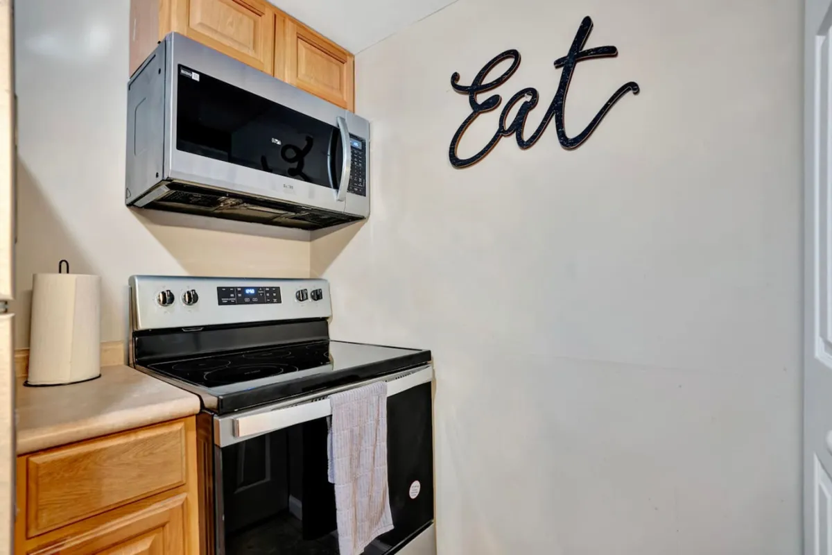 A bright and inviting kitchen space featuring wooden cabinets, a modern microwave and stove, and a decorative 'Eat' sign. The countertops are clear, emphasizing functionality and cleanliness, while the layout suggests ample room for meal prep and socializing.
