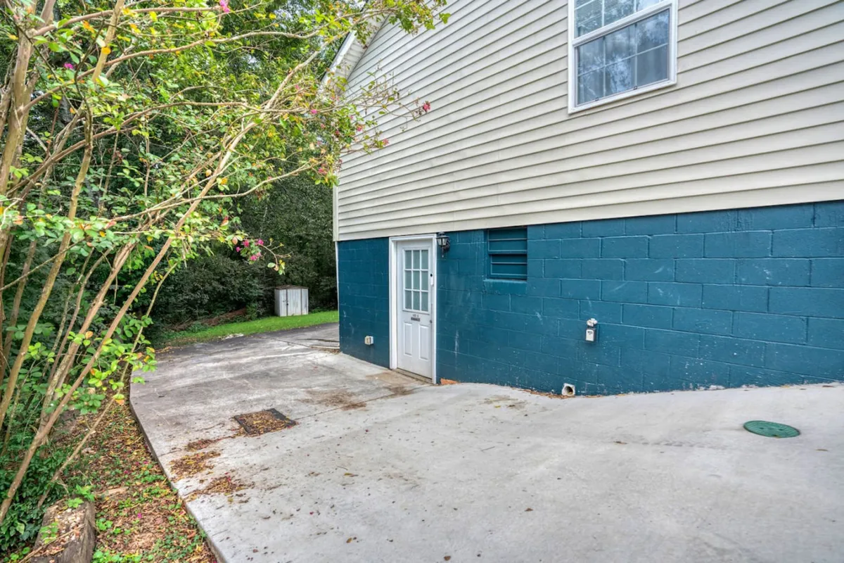 A charming exterior featuring a blue painted wall, a white door, and lush surrounding greenery. The space suggests areas for gardening, outdoor relaxation, and barbecuing, evoking a calm and inviting mood. The scene hints at a welcoming environment where families can enjoy quality time outdoors.