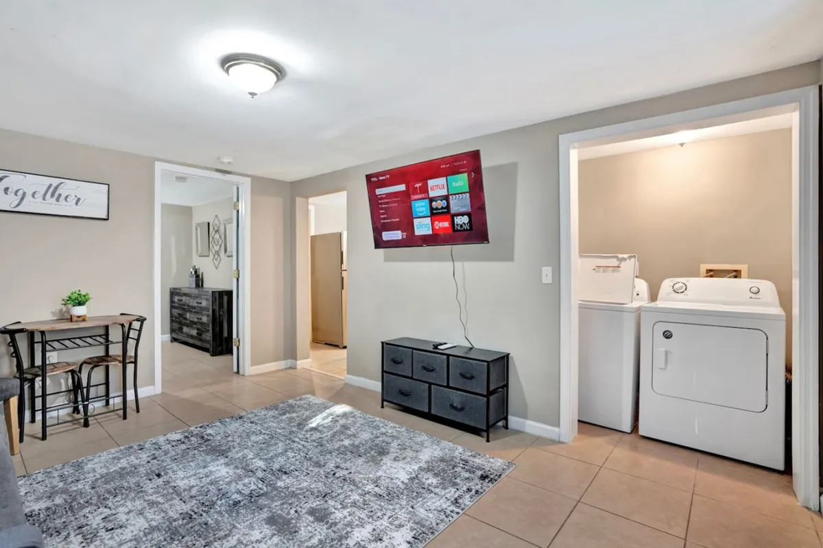 A view of the dining area featuring a dining table and chairs, adjacent to a mounted TV and a laundry area. The space is casual and comfortable, perfect for gathering, dining, or catching up over a meal.
